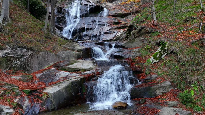 Majestic autumn waterfall cascading through a forest, surrounded by vibrant fallen leaves and smooth rocks in a peaceful, natural landscape. Aerial nature video.