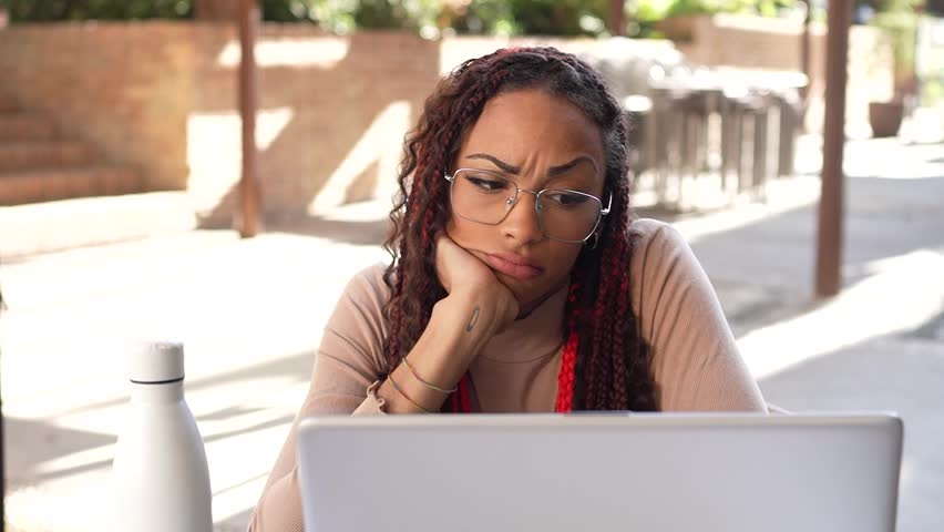 A woman with intricately braided hair gazes thoughtfully at her laptop while sitting in an outdoor setting, deeply reflecting on her work and the tasks she has ahead of her to accomplish
