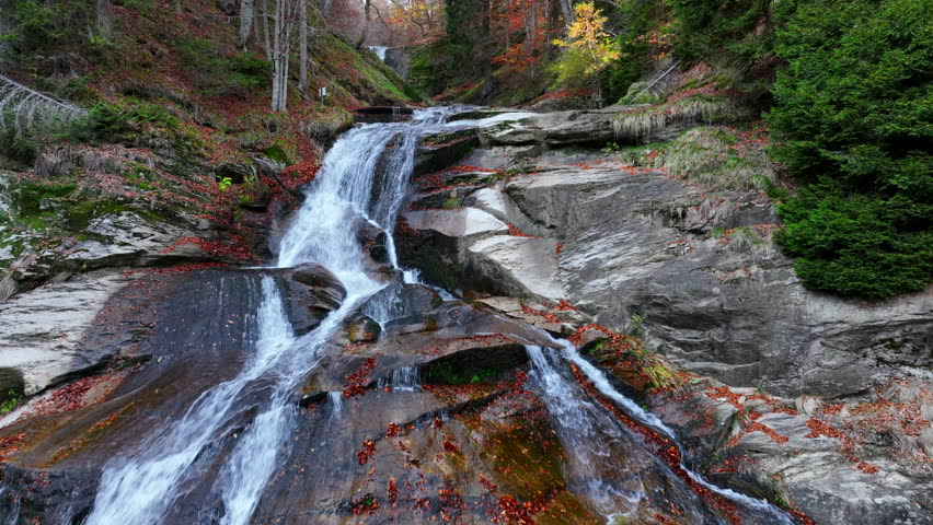 A stunning autumn forest waterfall, with silky water cascading over mossy rocks and vibrant fallen leaves in a serene natural environment. Aerial nature video.