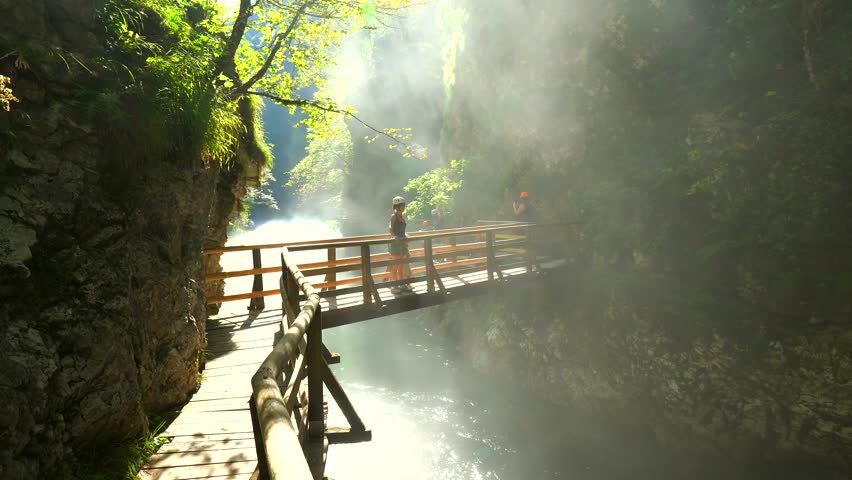 Sunbeams illuminating mist rising from radovna river in vintgar gorge near bled, with tourist walking on wooden footbridge