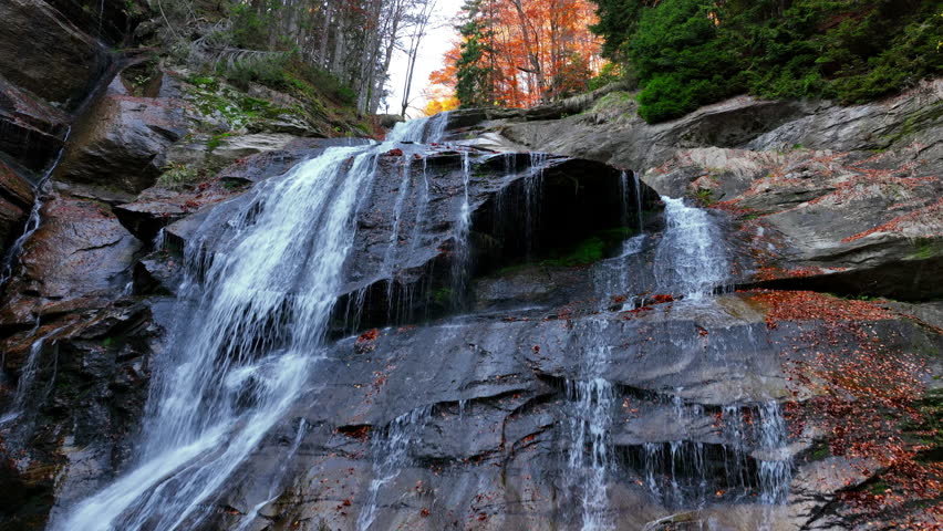 Silky waterfall flowing through an autumn forest, surrounded by fallen leaves and tranquil scenery, creating a harmonious natural landscape. Aerial nature video.