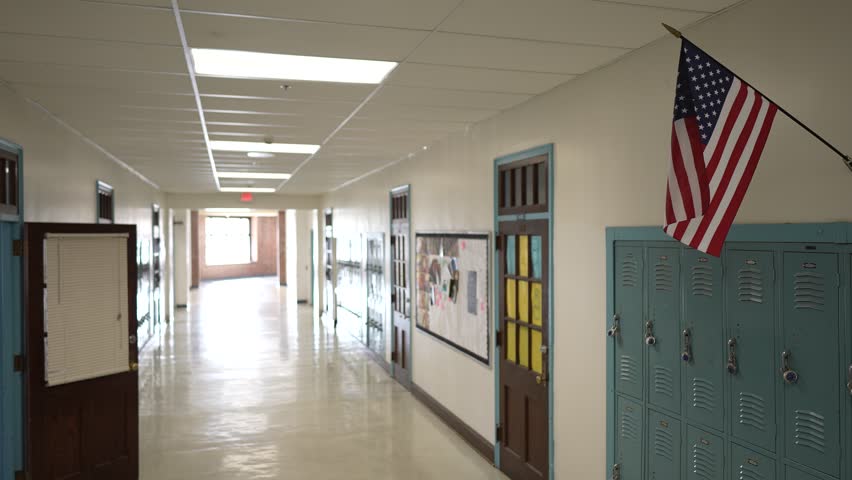 Brightly lit hallway in a school featuring rows of lockers and an American flag hanging above. The space reflects the calm before the day