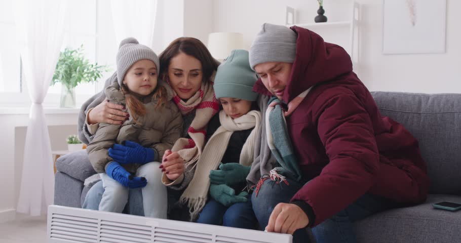 Parents, small children freezing at home wearing winter warm clothes sitting close near radiator heater, badly shivering with strong winter cold at home, low temperature problem, dissatisfied tenants