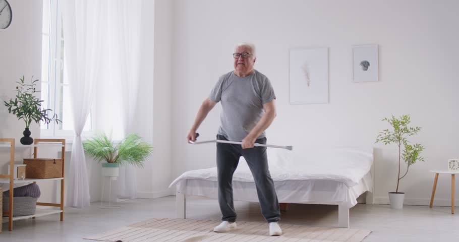 Happy excited senior elderly man dancing near bed holding crutch in bedroom. Joyful male person having fun at home with cane after successful treatment and rehabilitation. 4k video.