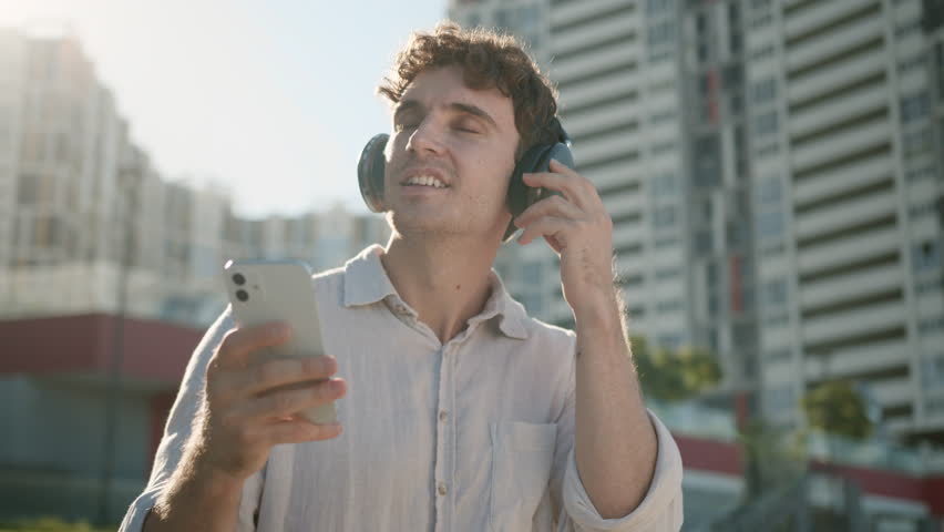 Happy Caucasian man smiling enjoyment listening music headphones earphones electronic gadget device technology favorite song guy businessman mobile phone smartphone modern business city male outside