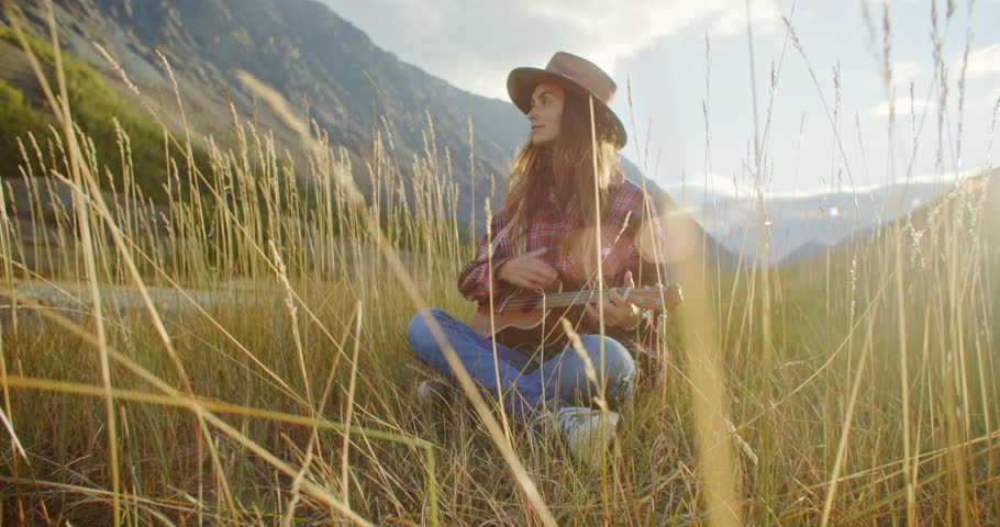 Young woman playing ukulele in a peaceful landscape with majestic mountains and tall grass. Female enjoying lovely music in a charming, serene outdoor setting