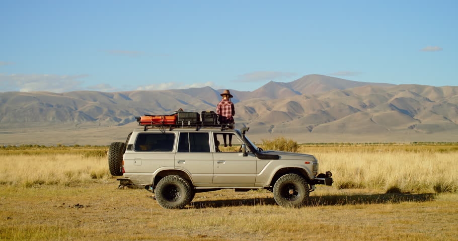 Young woman person stands proudly on an impressive offroad vehicle and take selfy on phone on roof a vehicle, surrounded by stunning mountains and open plains