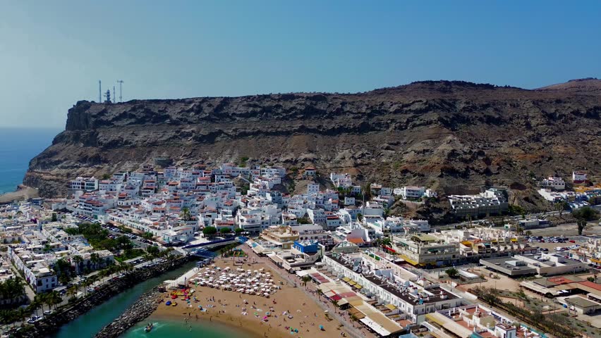 A small beach at the foot of a mountain with fine sand and clear blue water where leisure activities take place, is surrounded by steep volcanic cliffs and a quiet village with white houses.