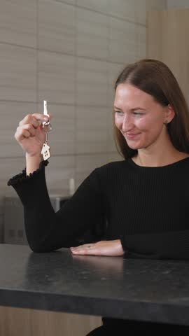 Woman holds new house key adorned with house-shaped keychain, smiling joyfully while gazing at her achievement of homeownership and excitement of new beginning. vertical video