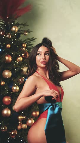 Sexy brunette in red lace bodysuit posing near Christmas tree. Gift satin bow tied around woman