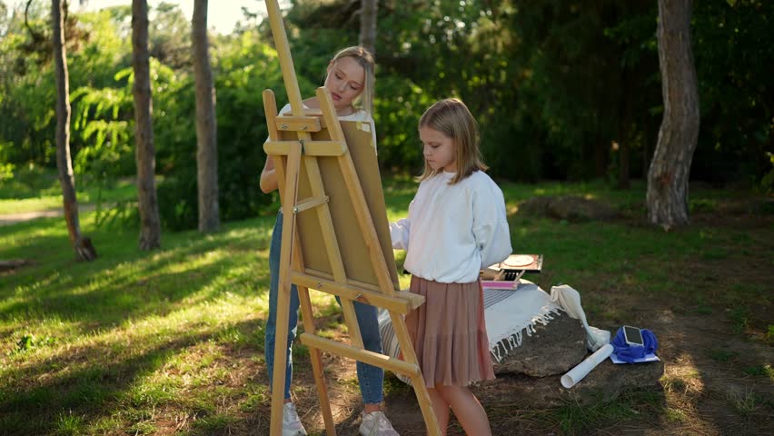 A young art teacher assisting a girl during a painting session on an easel in a tranquil park setting fostering creativity and learning