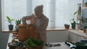 Medium shot of mature Caucasian woman taking fresh vegetables from brown paper bag while organizing meal preparation on wooden countertop in bright kitchen filled with green plants and sunlight - Powered by Shutterstock - Get 15% off with code: PIKWIZARD15