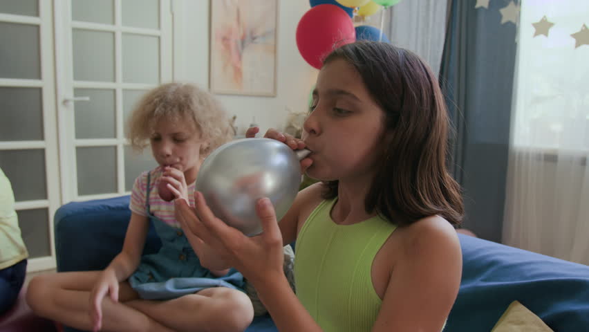 Girl blowing up silver balloon while getting ready for childrens birthday celebration, friend in background