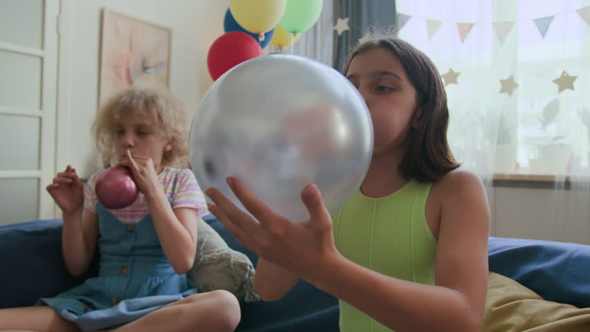 Girl blowing up silver balloon while getting ready for childrens birthday celebration, friend in background