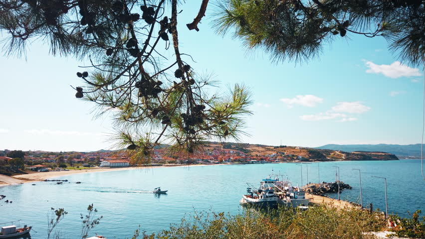 View of a greek town through fir branches. Pier, moored and mooving boat, beach, buildings, hills