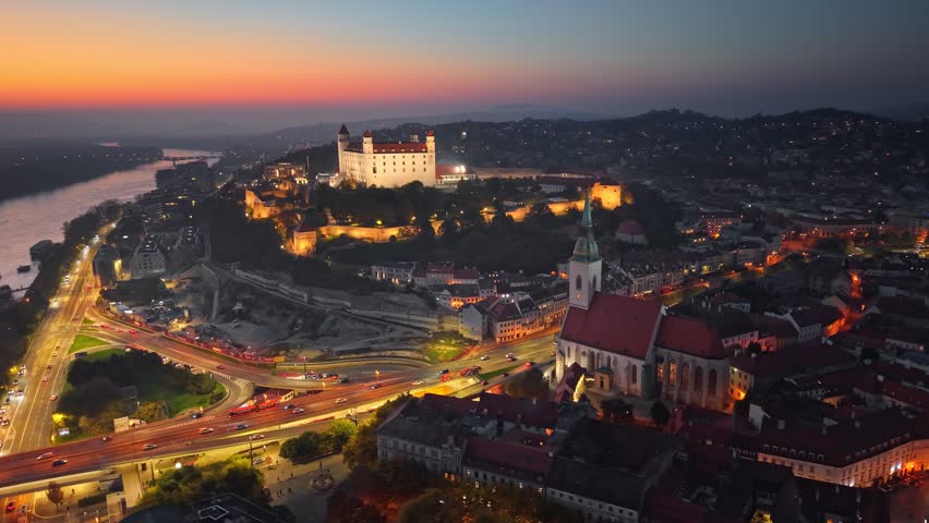 Sunset aerial view of Bratislava with illuminated castle and bustling city life along Danube River. Flying around St Martin