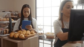 Woman working in a bakery using a tablet while another woman operates the register, surrounded by fresh pastries in a cozy indoor shop setting - Powered by Shutterstock - Get 15% off with code: PIKWIZARD15