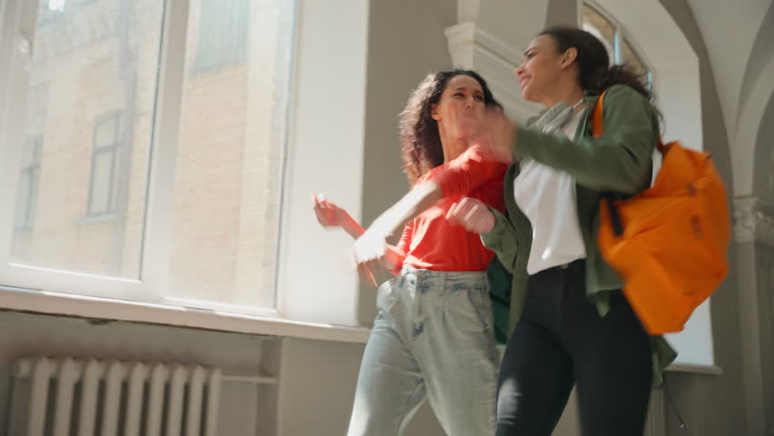 Two multiracial women friends multiethnic African American girls female diversity Hispanic Latina students together walking dancing going in university college academy corridor hall good carefree mood