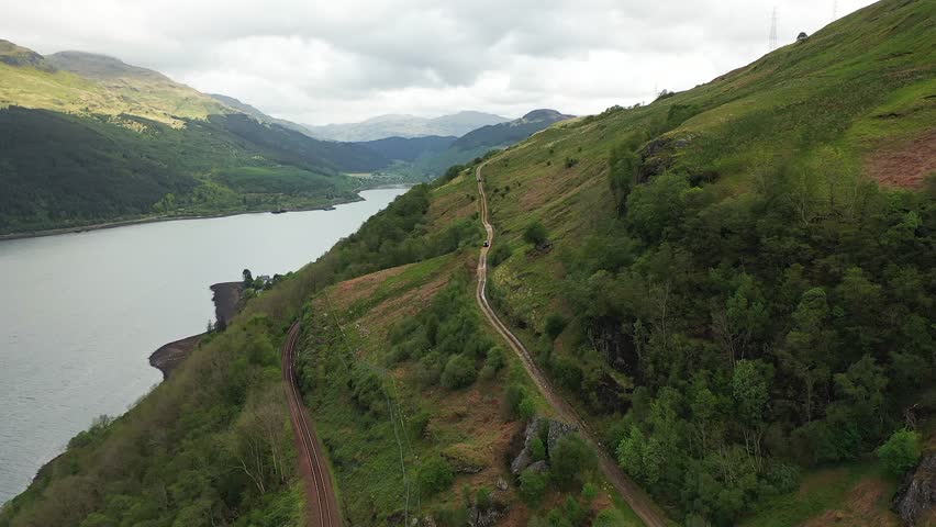 Aerial above dirt track and railway track on mountain slope above Loch Long in beautiful valley, Scottish Highlands, Scotland, United Kingdom.