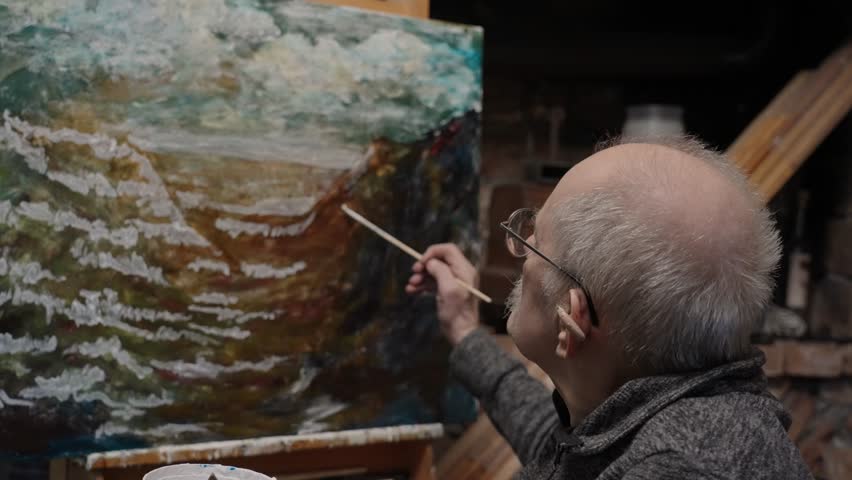 Senior artist on a wheelchair paints an oil painting on canvas in a studio