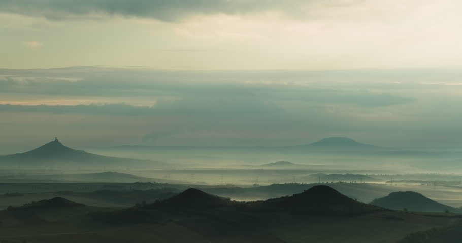 Timelapse of hilly landscape with a silhouette of castle ruins on one hillwith mist rolling in valleys and shadows transforming with time and of overcast sky white and grey clouds on a cloudy day 