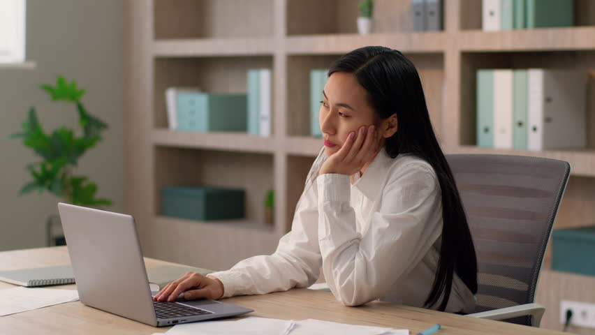 Dreamy Asian Chinese business woman female Japanese Korean girl businesswoman office worker secretary manager employee working typing laptop pc looking distance daydreaming thinking dreaming indoors