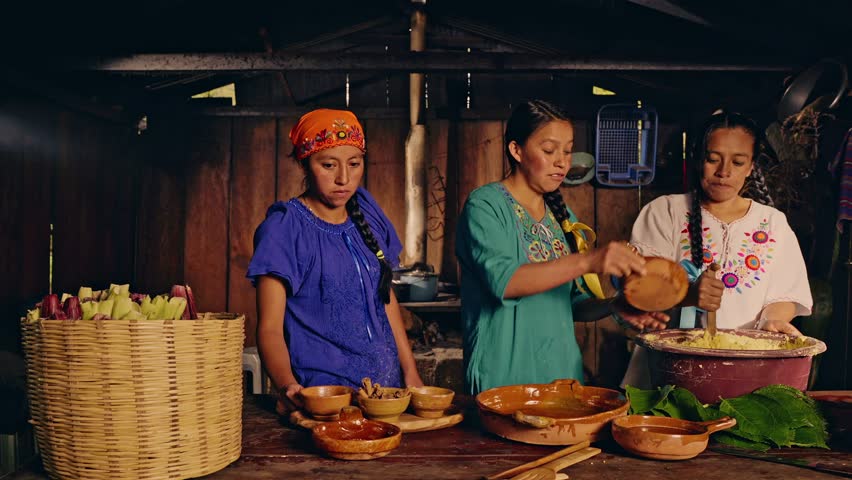 Three Latin women from the Xinca ethnic group work in front of a wooden table and make tamales by mixing ingredients.	