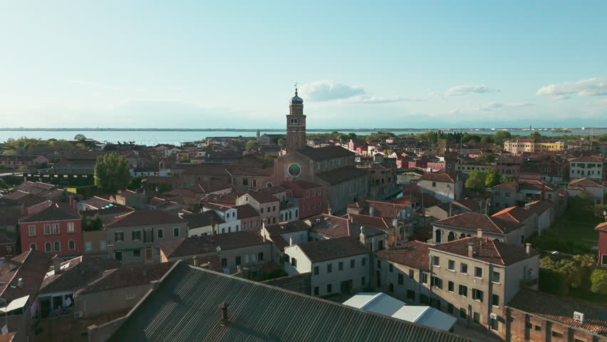Aerial footage of a historic church and canal in Murano, Venice, with scenic views of the surrounding cityscape and lagoon on a sunny day.