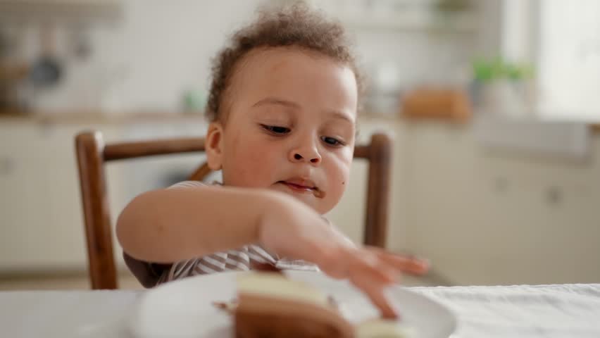 Baby boy eating chocolate cake in family dinner or Birthday party, dirty face. Happy childhood, dietic food for toddlers, mixed race little child sitting at table in home kitchen, slow motion shot