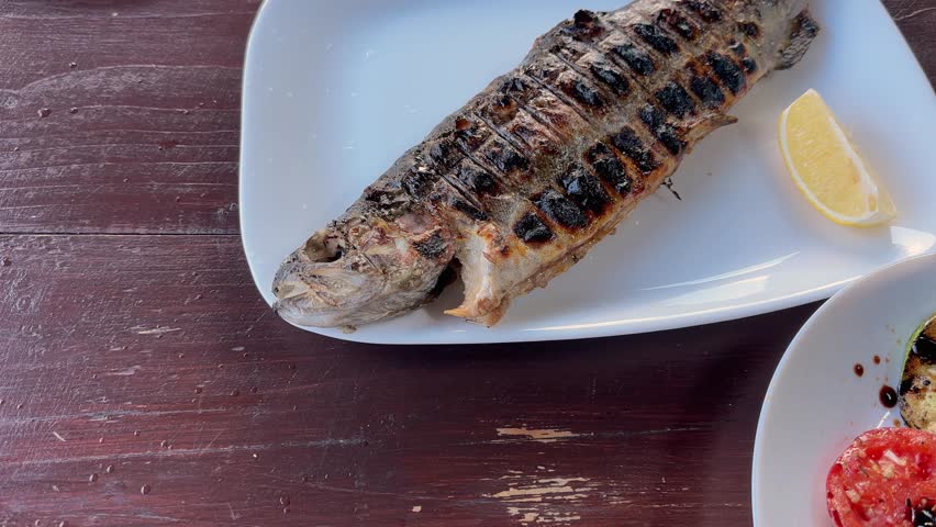 Whole grilled river trout with fresh lemon slices on the white dish on a wooden table outdoors, view close-up while panning with rotation
