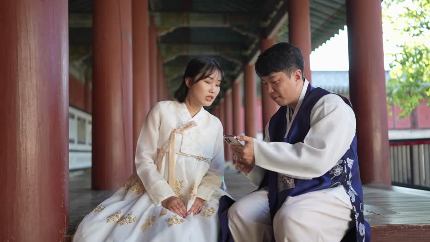 A Korean couple, a man in his 30s and a woman in her 20s, wearing hanbok from Seoul, South Korea, are sitting in a historical building, drinking water and using their smartphones. Slow motion video.