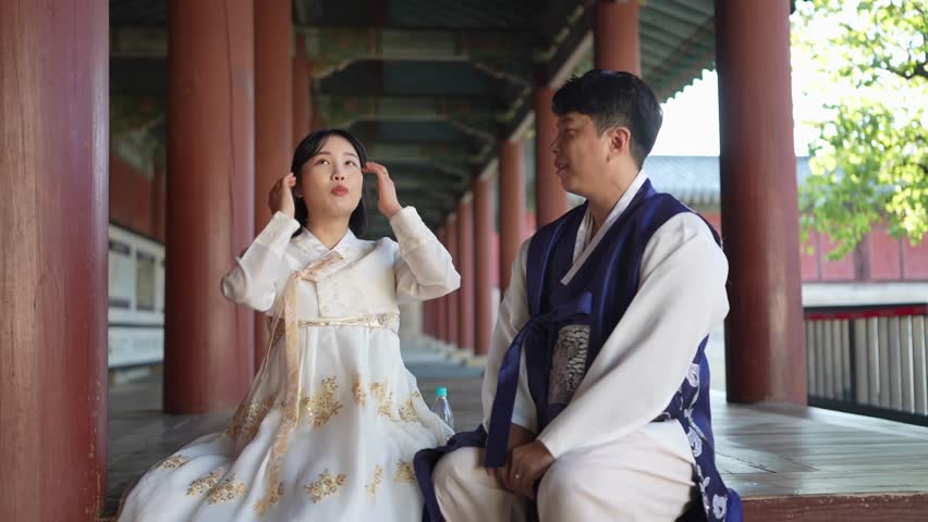 A Korean couple, a man in his 30s and a woman in her 20s, both from Seoul, South Korea, wearing Hanbok, are sitting side by side in a historical building and talking. Slow motion video.