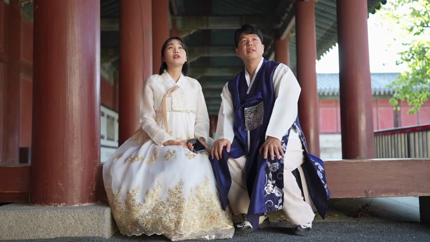 A Korean couple, a man in his 30s and a woman in her 20s, both from Seoul, South Korea, wearing Hanbok, are sitting side by side in a historical building and talking. Slow motion video.