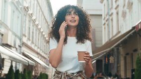 Front camera view of cute Caucasian woman with curly hair walking in middle of street. Talking on phone with husband or relative. Smiling during conversation. Sipping hot drink from small cup. - Powered by Shutterstock - Get 15% off with code: PIKWIZARD15