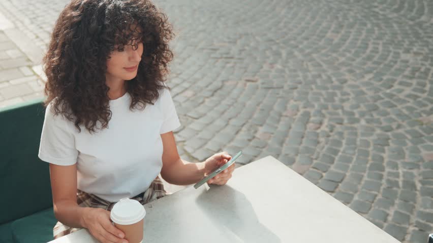 View from above of Caucasian female sitting at table. Paying for her order in restaurant with phone. Completing transaction by lowering smartphone to payment terminal. Modern technology concept.