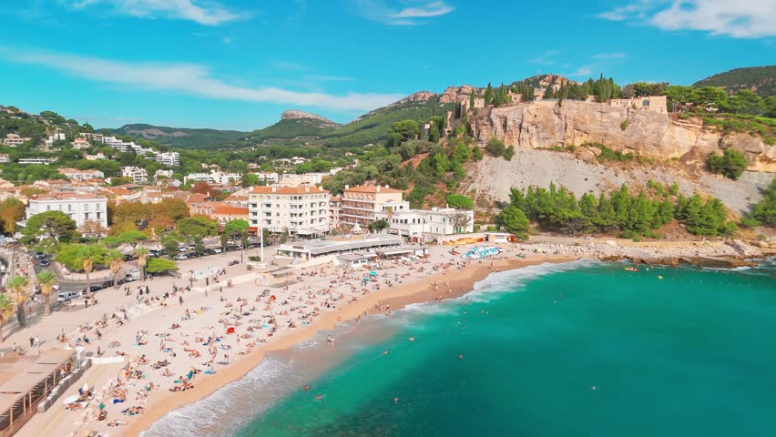 Aerial view of Cassis on the French Riviera, southern France. The scene features a medieval castle perched on a cliff, a sandy beach, turquoise sea waters, and the historic buildings of the town