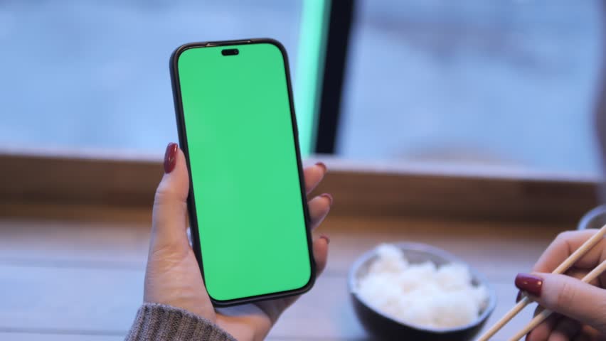 A hand holding a modern smartphone featuring a green screen, perfect for creating app mockups in a cozy cafe