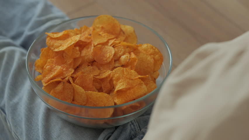 Girl Eating Chips From Bowl While Sitting on Floor in Living Room. Female Eating Unhealthy Potatoes Crisps. Snack on Junk Food. Child Eats Fast Food, Junk Food at Home Watching TV. Childhood Obesity.