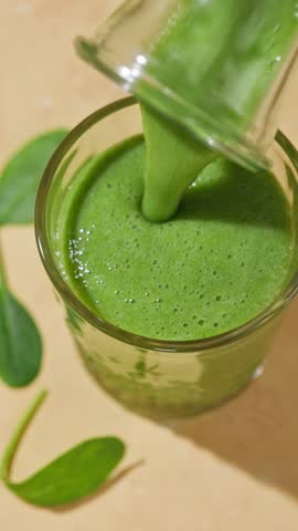 freshly made green spinach and banana smoothie flows into a glass from a bottle on a sunny light table surface