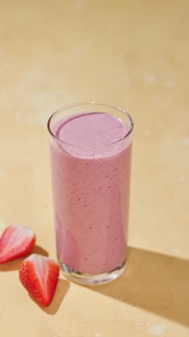 hand puts a straw into a glass of freshly made pink strawberry and banana smoothie on a sunny light table surface