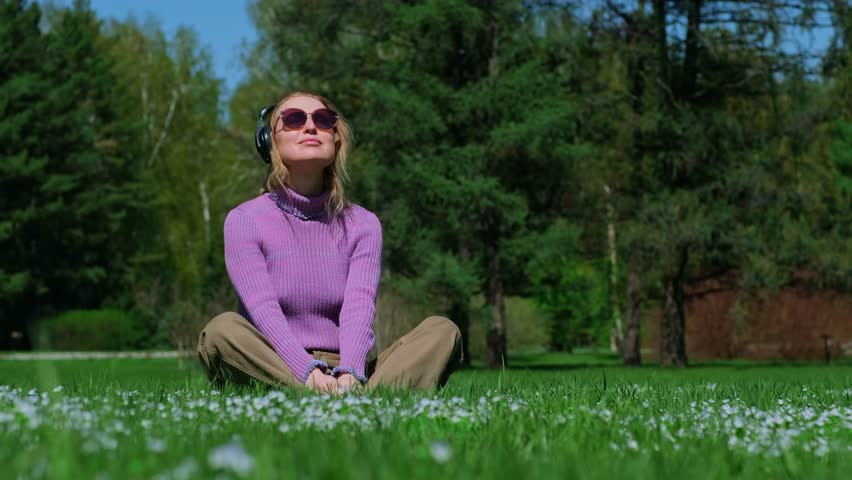 Happy adult woman listening to music with headphones while sitting on a green blooming lawn in a summer sunny park.