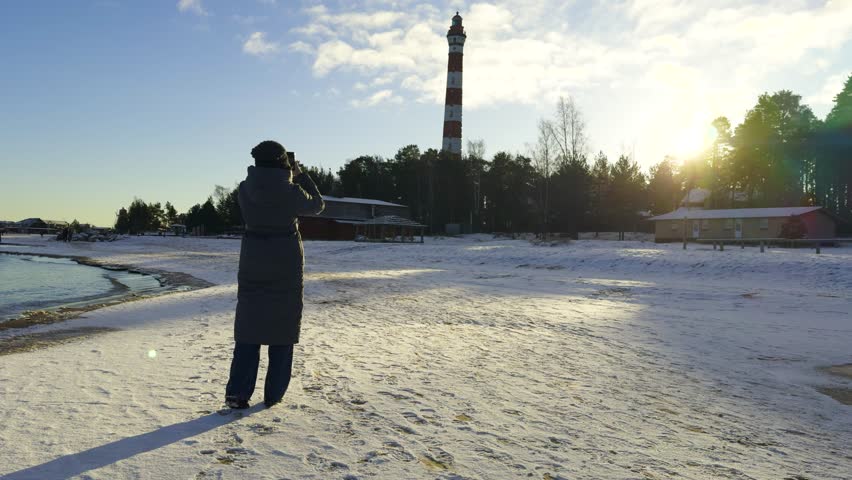 A girl in a long winter coat takes a photo of Osinovets Lighthouse on the snowy shore of Lake Ladoga during sunset. The sun casts long shadows, with a forest and houses in the background