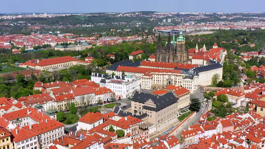 A breathtaking view of Prague showcases the iconic castle, red rooftops, and lush green hills under a bright blue sky, capturing the city
