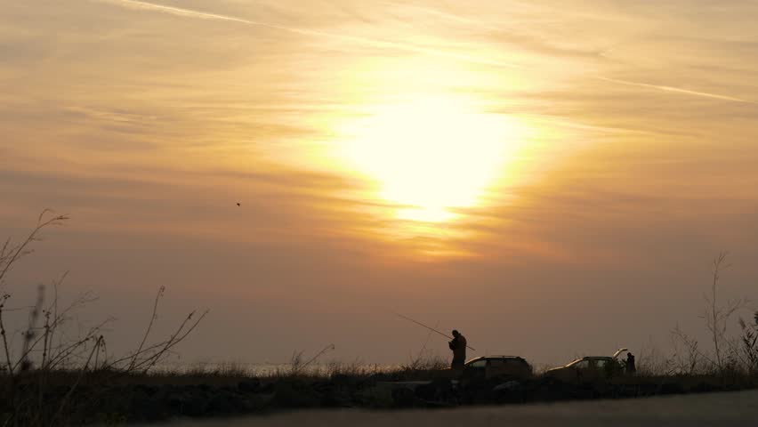 Unrecognizable men with fishing rod on the edge of shore in bay with coastline in background. Fishermen fishing outdoors in sea during sunrise in golden hour, copy space