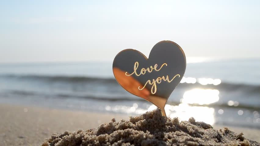 Plastic stick shape of heart with words Love you in sand beach of sea shore on background sea waves on sunny summer day close-up. Concept love relationship romance amour celebration St Valentines Day