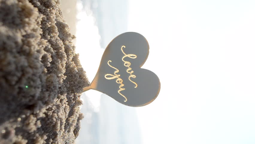 Plastic stick shape of heart with words Love you in sand beach of sea shore with sea waves on sunny summer day close-up. Concept love relationship romance amour celebration St Valentines Day Vertical