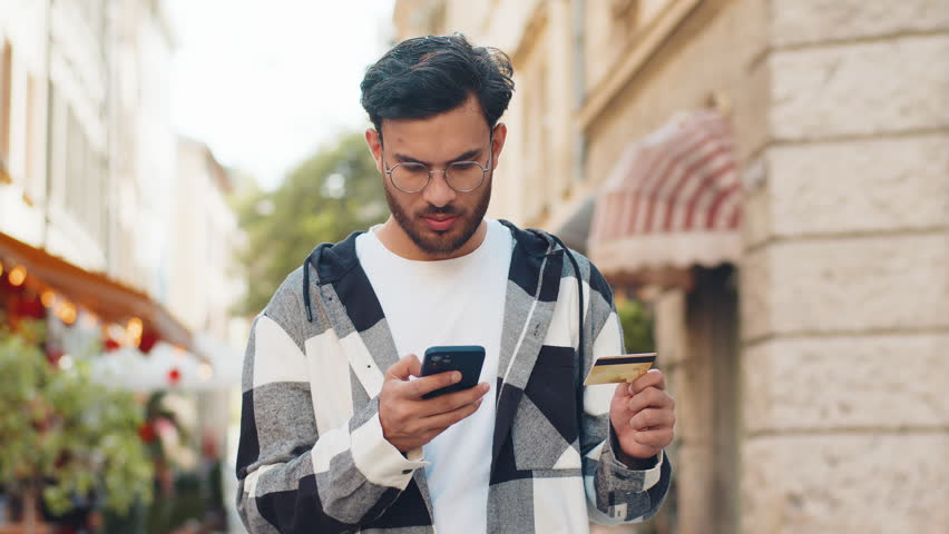 Shocked Indian man trying to pay online shopping with smartphone blocked credit card. Annoyed young Arabian tourist on street buying bank refuse problem unsuccessful payment lack of money balance.