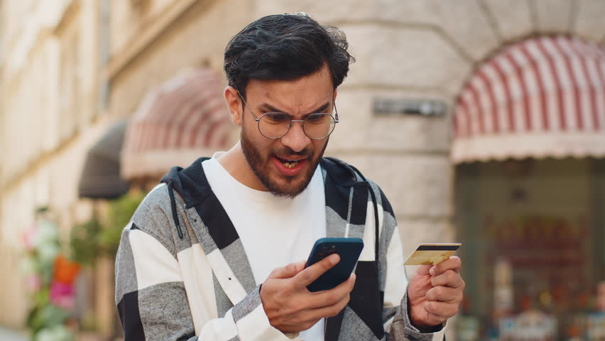 Shocked Indian man trying to pay online shopping with smartphone blocked credit card. Annoyed young Arabian tourist on street buying bank refuse problem unsuccessful payment lack of money balance.