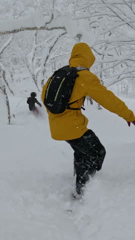 VERTICAL: Two snowboarders carve through Rusutsu’s deep powder snow, weaving between snow laden trees in a serene, dense forest. The untouched terrain and fresh lines offer an epic winter adventure.