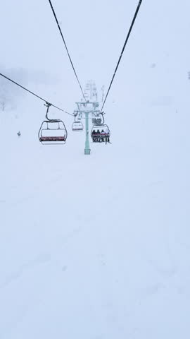 POV, AERIAL: Ski lift ride through heavy snowfall in Niseko, Japan. Skiers and snowboarders carve fresh powder below, navigating rugged terrain and whiteout conditions amid an intense winter storm